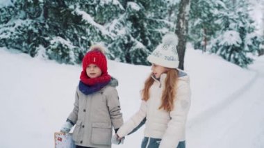 Beyaz şapkalı tatlı bir kız ve kırmızı şapkalı bir çocuk kışın köknar ağaçlarıyla çevrili yolun ortasında yürüyen ve mühürlü bir zarfa bakıyorlar. Yakın plan. Yavaş çekim