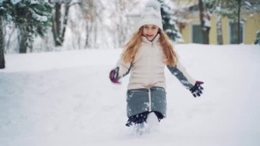 Sıcak elbiseli küçük bir kız tepeden iniyor ve kara dokunuyor. Çocuk parkta kar yağışının arka planına elleriyle kar saçıyor. Yavaş çekim