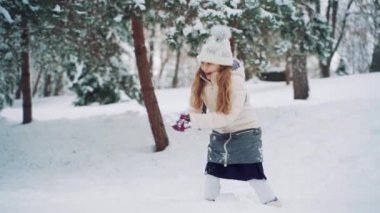 Beyaz ceketli ve mor eldivenli neşeli kız kışın köknar ağaçlarının arka planında parka kartopu atıyor. Yavaş çekim