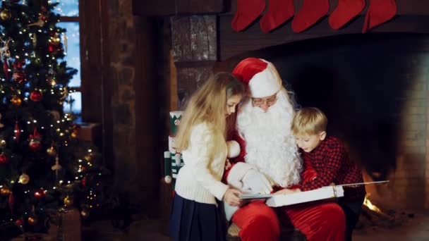 圣诞老人正在向孩子们展示一本印有照片的相册 相册的背景是一个有圣诞袜的壁炉 房间里有一棵圣诞树 但穿着格子呢衬衫的男孩却跑掉了 — 图库视频影像
