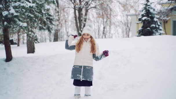 暖かいダウンジャケットと帽子の美しさの女の子は彼女の手の中に雪玉を保持し 家の近くの丘の冬の寒い天候でそれを前方にスローします スローモーション — ストック動画