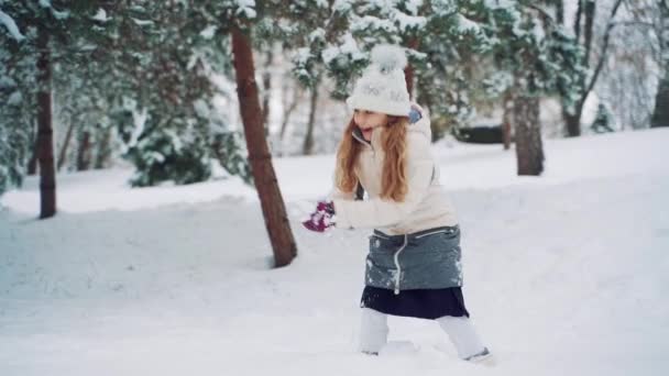 白いダウンジャケットと紫色の手袋の陽気な女の子は 冬のモミの木の背景に公園で雪玉を投げています スローモーション — ストック動画