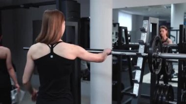 Atletik genç kadın ağırlık barına doğru yürür ve onu aynanın karşısındaki spor salonunda kaldırır. Dayanıklılık egzersizi