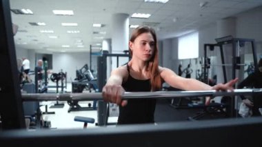 Siyah bluzlu ve dalgalı saçlı güçlü bir kadın spor salonundaki ağırlık çıtasını yükseltiyor. Yakın plan.
