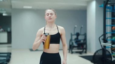 Siyah bluzlu güzel bir kadın sarı bir içecekten içiyor ve spor malzemelerinin arka planında spor salonunda yürüyor. Sağlıklı yaşam tarzı