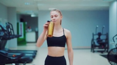 Siyah spor giysili genç bir kadın elinde bir sallayıcıyla spor salonuna giriyor ve ondan su içiyor. Spor salonu. Bulanık Arkaplan