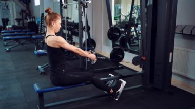 Spor taytlı ve üstteki ince topuzlu kadın spor salonundaki spor ekipmanlarının arka planında kürek simülatörü üzerinde egzersiz yapıyor..