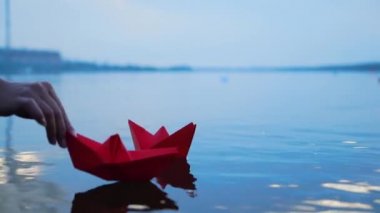 Kadının eli suya kağıttan tekne koyuyor. Güzel yaz günbatımı. Kağıt tekne. Origami.
