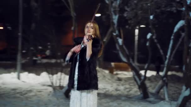 一个身穿白衣 化着妆的漂亮姑娘正在雪地的冬季公园里拉小提琴 相机向左运动 — 图库视频影像