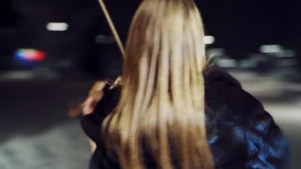 黒の半丈のコートを着た楽しい女の子がバイオリンを手に持ち 街の中心部の広場で冬の夜にそれを演奏しています 背景がぼやけている 周囲へのカメラの動き — ストック動画