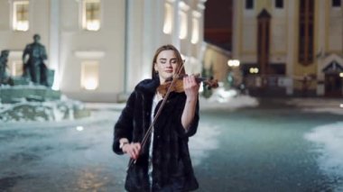 Gece elbisesi ve paltosu giymiş bir müzik fakültesi öğrencisi, arkasındaki anıt ve şehir binalarının arka planında kemanla ünlü bir müzik çalıyor. Yakın plan.