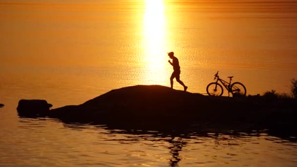 男性サイクリストは川の近くで休んでいます 川の近くに立って遠くの夕日を見ている若いアクティブなスポーツマンと彼のマウンテンバイクのシルエット — ストック動画