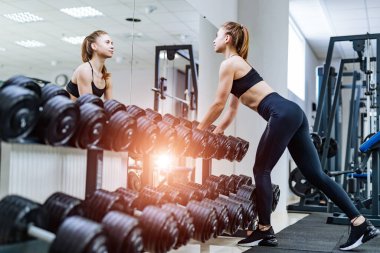 Metal halterlerle rafın yanında poz veren genç ve formda bir kızın tam boy portresi. Şirin, ince kadın başlık takıyor ve spor giysisi giyiyor. Duvardaki kameraya ayna yansıması ile bak.