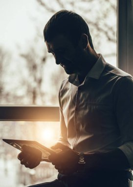 Elinde tabletle genç bir iş adamı. Genç bir uzmanın silueti. Büyük pencerelerin arkasındaki panoramik manzara. Serbest meslek kavramı. Akşam işi..