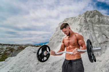 Kaslı ve sakallı bir adam dışarıda halterle egzersiz yapıyor. Vücut geliştirme ve açık hava sporları konsepti. Taş ocağında fotoğraf çekimi..