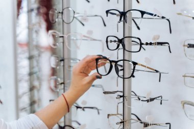 Choosing glasses. Client in optics. Selective focus on hand. Woman touching corrective glasses on stand with spectacles. Wide assortment in shop. clipart