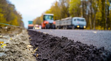 Yeni yol inşaatında çalışan yol makinelerinin geniş bir manzarası var. Önde taze asfalt var. Arka planda bulanık makineler.