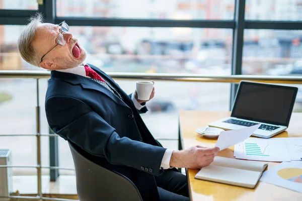 Tired mature business man with opened mouth and tongue out sitting in chair at table in modern office. Successful senior businessman sitting in meeting room with documents. funny man in suit in elegant office.