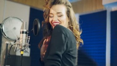 Harika bir kadın kayıt stüdyosunun arka planında profesyonel bir mikrofonun önünde neşeyle dans edip şarkı söylüyor. Mutlu müzisyen mikrofonun başında duruyor ve yeni bir müzik yapıyor.