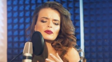 Siyah dalgalı saçlı, çıplak omuzlu çekici şarkıcı mavi arka planda mikrofona şarkı söylüyor. Kayıt stüdyosunda güzel bir kadın. Yakın plan.