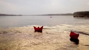 Kadının eli, günbatımında yüzen diğerleri için kırmızı kağıt tekneyi suya koyuyor. Origami akşamları nehirde dalgalanan gemiler.