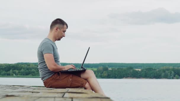 Fiatal férfi pólóban és barna rövidnadrágban ül a folyó mellett, és laptopot használ természetes háttérrel. Az ember befejezi a munkáját a laptopon, és élvezi a pillanatot, ül a fa híd.