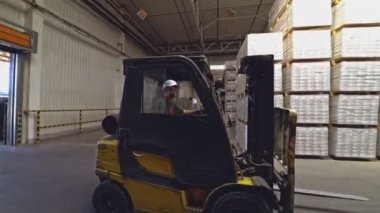 Kasklı bir operatör, büyük rafların arkasındaki doğru ürünü bulmak için depodan geçen sarı elektrikli bir forklift kullanıyor..