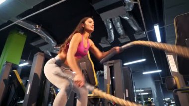 Uzun siyah saçlı güçlü kız trisepsleri spor malzemelerinin arka planındaki kabloları sallayarak eğitiyor. Spor kıyafetli mutlu genç kadın içeride egzersiz yapıyor. Yakın plan.