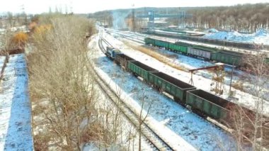 Konteynırları olan bir yük treni bir kış günü raylar boyunca şehre doğru hareket eder. Malların nakliyesi. Hava görünümü.