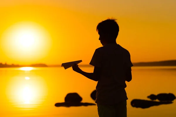 Schwarze Silhouette eines Jungen, der vor orangefarbenem Sommersonnenuntergang steht. mit einem kleinen Papierflieger in der Hand. Horizontales Foto. — Stockfoto