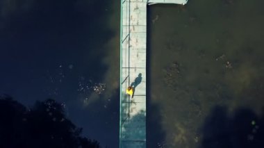 Küçük çocuk uzun tahta köprüde yürüyor ve güneşli bir günde suya atlıyor. Yelek giyen çocuk yaz tatilinde nehirde iyi vakit geçiriyor. Hava görünümü