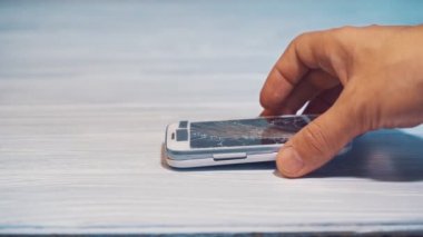 Erkek eli gri ahşap yüzeydeki çatlamış beyaz akıllı telefona dokunuyor. Bir adamın elinde kırık bir cep telefonu. Yakın plan.