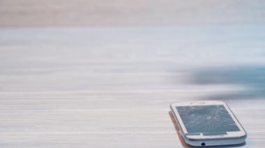 Bir adamın elinin ağır çekimde yerden kırık bir akıllı telefonu alması. Pahalı bir cep telefonunun kırık ekranı içerideki ahşap zeminde yatıyor..