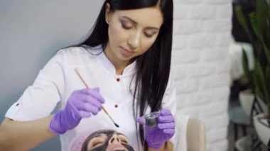 Deneyimli bir kozmetikçi güzellik salonundaki bir kadının yüzüne siyah temizleyici maske takar. Yakın plan. Güzellik endüstrisi.