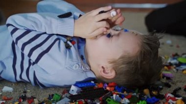 Küçük çocuk yere uzanmış ve elleri üzerine düşen lego parçalarından saklanarak yüzünü kapatıyor. Çocuğun yüzüne plastik renkli tuğlalar dökülüyor. Yakın plan.