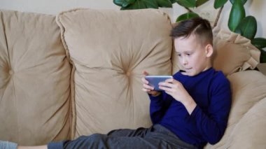 Akıllı telefonuyla oynayan genç çocuk sinirleniyor ve eve alet fırlatıyor. Rahat giyimli gergin çocuk kanepede oturup telefonda oyun oynuyor..