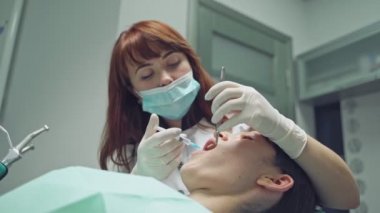 Klinikte oral ameliyattan önce kadın stomatolog anestezi uyguluyor. Güzel dişçi işi sırasında ağız aynası ve ilaç şırıngası kullanıyor. Yakın plan.