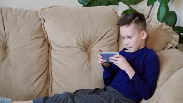 スマホで遊んでいる10代の少年は怒って家にガジェットを投げるようになります ソファに座って電話でゲームをしているカジュアルな服の神経質な子供 — ストック動画