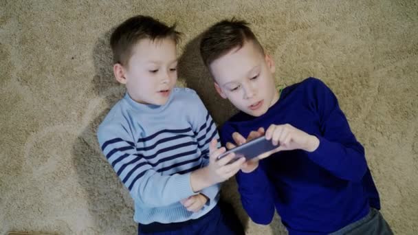 2人の幸せな男の子が床に横たわって 一緒に1台のスマートフォンで遊んでいます リラックスした子供たちは 柔らかい敷物の上で現代的なガジェットと完全に楽しんでいます — ストック動画