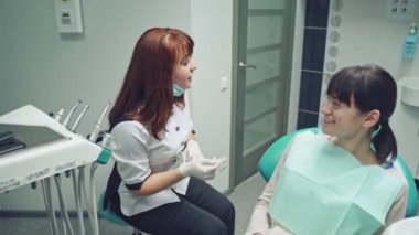 Kadın doktor, dişçi odasının arka planındaki dişlere nasıl bakacağını hastasına açıklıyor. Uzman, stomatoloji ofisindeki tıbbi sandalyede oturan hastaya beş dakika veriyor..