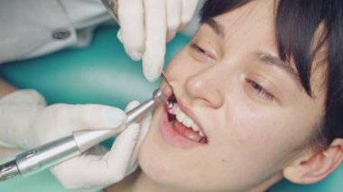 Genç bir kadın kozmetik diş hekimliğinde profesyonel diş temizliği alıyor. Hastanın yüzü ve stomatoloğu dişçi muayenehanesinde küçük fırçaları olan bir aletle çalışıyor. Yakın plan.