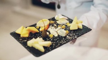 Bir garson elinde farklı şekillerde sert peynirle süslenmiş ve restorana susam serpiştirilmiş koyu ahşap bir tahta tutuyor. Peynir tatma.