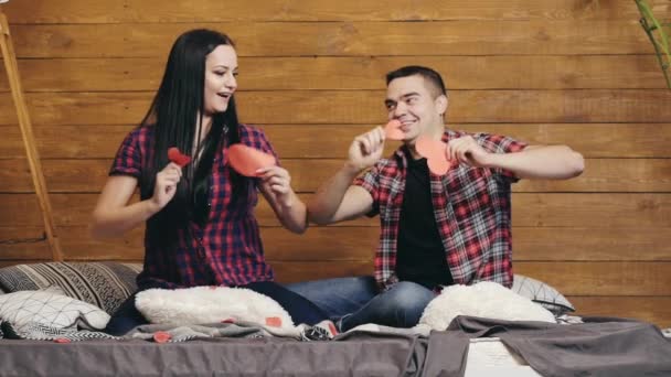 両手で赤い紙の心を持つベッドの上に座っているロマンチックなカップルは手で動きます 幸せな男は手のさまざまな側面に動きを示し 彼の女の子は彼の後に繰り返されます — ストック動画