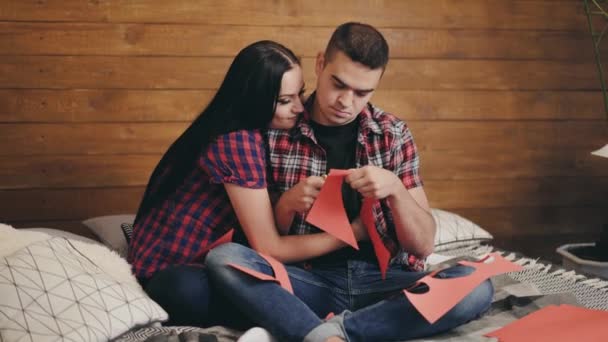 英俊的男人正在木制背景下从红纸上割下心脏给他的女朋友 这对快乐的夫妇坐在床上 女孩拥抱着并亲吻着她的男朋友 — 图库视频影像