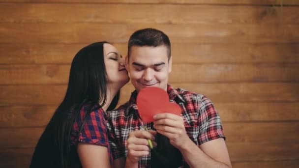 長い髪の女性は彼女のボーイフレンドといちゃつくと 彼は木製の背景に隔離された紙の赤いハートを彫っています 若いカップルの愛一緒に時間を過ごす バレンタインデー — ストック動画