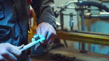 Tamirci bir atölyede metalle çalışıyor, metal parçalarının ölçümlerini yapıyor. İşçi elinde bir kalibre tutuyor, fabrika arka planında özel bir takım elbise..
