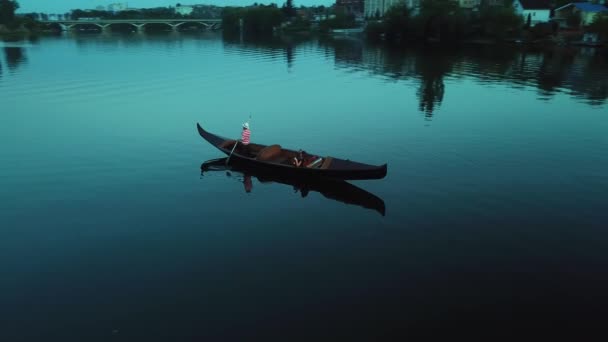 夜には川の背景にボートの中で Oarでボートを漕ぐ帽子の強い男の空中ビュー 女性はゴンドラに座って夜の美しい風景を楽しむ — ストック動画