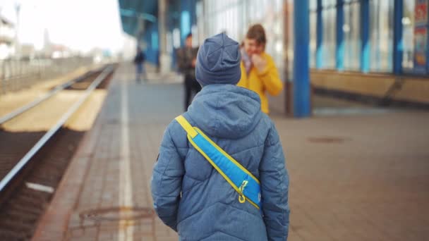 ホームに一人で立って電車を待っている少年の裏側のビュー 灰色の帽子の男の子と鉄道駅のバックパック付きのジャケット — ストック動画