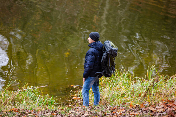 Путешественник с рюкзаком стоит на берегу реки с лазурной водой в солнечный день осенью. Путешествие. Пейзаж с подходящим человеком, апельсиновые деревья осенью. Пешие прогулки. Вид сзади

