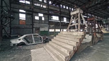 Merdivenleri ve kırık bitkinin arka planında antika araba çerçevesi olan eski bir yapı. İçinde hasarlı bir araba ve metal döküntüleri olan terk edilmiş büyük bir binanın içinde..